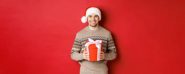 Porträtt av stilig man som håller en present, önskar glad semester, står i tomte hatt och jul tröja mot röd bakgrund — Stockfoto