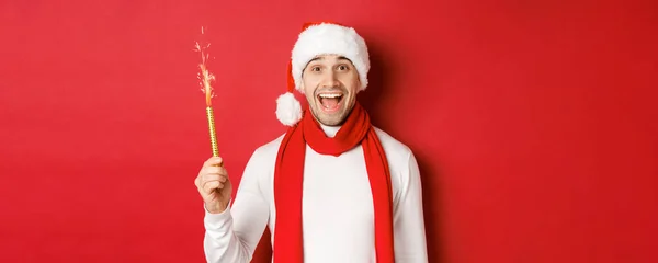 Koncepcja Bożego Narodzenia, ferie zimowe i uroczystości. Przystojny mężczyzna świętuje Nowy Rok i dobrze się bawi, trzyma iskrę i uśmiecha się, nosi czapkę Świętego Mikołaja, stoi na czerwonym tle — Zdjęcie stockowe