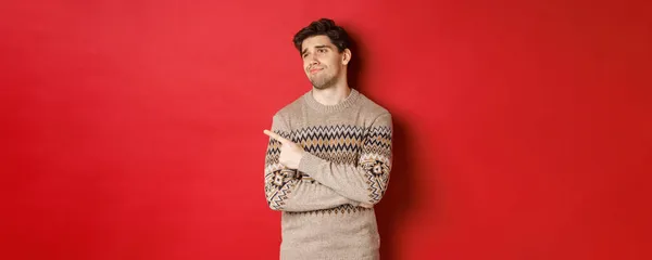 Образ розчарованого красивого чоловіка в різдвяному светрі, дивлячись і вказуючи ліворуч з засмученим обличчям, висловлює жаль, показуючи новорічну рекламу, стоїть на червоному тлі — стокове фото