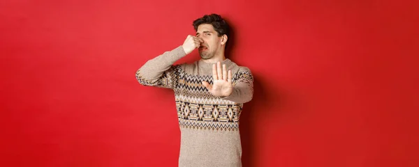 Obrázek znechuceného pohledného muže v zimním svetru, jak odmítá něco se špatným zápachem, zavírá nos a zvedá ruku, stojí nad červeným pozadím — Stock fotografie