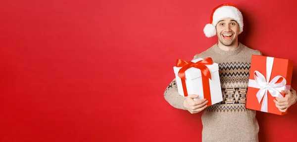Imagem de cara atraente alegre segurando presentes de Natal, de pé em chapéu de santa e suéter de inverno, sorrindo feliz, de pé sobre fundo vermelho — Fotografia de Stock