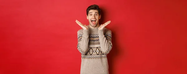 Retrato de cara surpreso e animado em camisola de Natal, reagindo ao presente de ano novo, sorrindo espantado, de pé sobre fundo vermelho — Fotografia de Stock