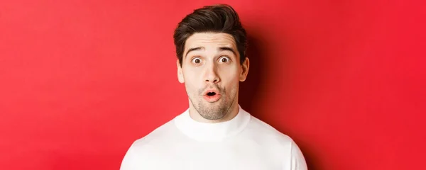 Close-up of surprised handsome man in white sweater, open mouth and looking wondering, hear amazing news, standing over red background — Stok Foto