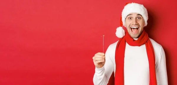 Koncepcja Bożego Narodzenia, ferie zimowe i uroczystości. Zbliżenie szczęśliwego przystojniaka w czapce i szaliku Mikołaja, bawiącego się na noworocznej imprezie, trzymającego iskrę i uśmiechnięte, czerwone tło — Zdjęcie stockowe