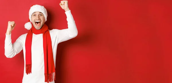 Образ радостного кавказца в шляпе и шарфе Санты, кричащего от радости и поднимающего руки, празднующего победу или победу, торжествующего над красным фоном — стоковое фото