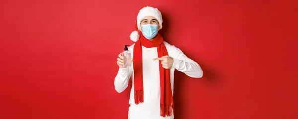 세계적으로 유행하는 시기에는 covid-19, 크리스마스, 그리고 휴일의 개념이다. 산타 모자와 의료용 마스크를 쓴 잘생긴 행복 한 남자, 빨간 배경 위에 서 있는 손 소독 기를 추천 — 스톡 사진
