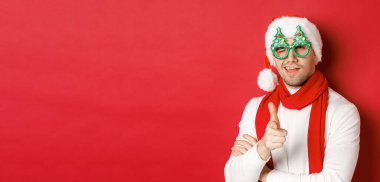 Noel, kış tatili ve kutlama kavramı. Noel Baba şapkası ve parti gözlüğü takmış, gülümseyen ve kameraya silah doğrultan, kırmızı arka planda duran arsız genç adamın yakın çekimi.