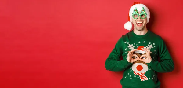 Portret beztroskiego przystojniaka w czapce Mikołaja i okularach imprezowych, śmiejącego się ze swojego świątecznego swetra, szczęśliwie wyglądającego na czerwonym tle — Zdjęcie stockowe