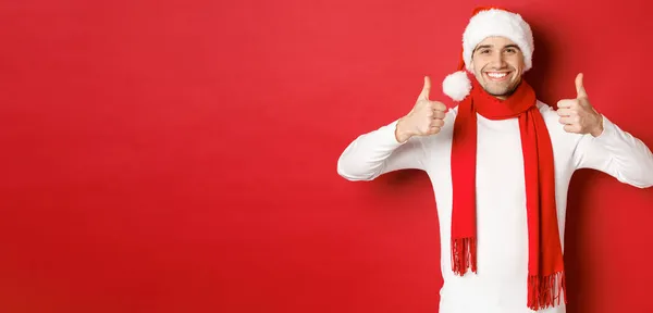 Πορτρέτο του όμορφου χαμογελαστού άνδρα με το κασκόλ και το καπέλο, που δείχνει τους αντίχειρες, γιορτάζει τα Χριστούγεννα, στέκεται πάνω από το κόκκινο φόντο — Φωτογραφία Αρχείου