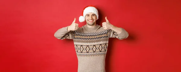サンタの帽子とセーターの魅力的な幸せな男の肖像、承認と笑顔で親指アップを示し、陽気なクリスマスを望み、赤い背景の上に立って — ストック写真