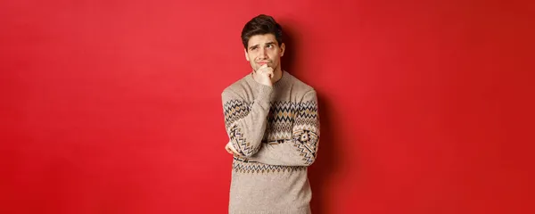 Portrait of confused man in winter sweater, thinking about christmas presents, looking troubled, searching for good gifts, standing over red background — Stock Photo, Image