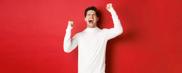 白いセーターのハンサムな男の肖像、陽気で、勝利を祝い、喜びを叫び、勝利に手を上げ、赤い背景の上に立つ — ストック写真