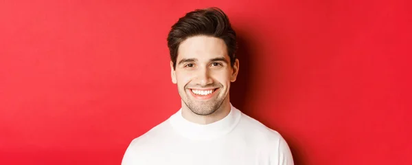 Lähikuva komea hymyilevä brunette mies, yllään valkoinen pusero, hymyilevä onnellinen ja luottavainen, seisoo punaista taustaa vasten — kuvapankkivalokuva