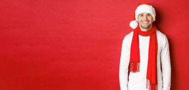 Mutlu beyaz adam Noel 'i kutluyor ve gülümsüyor, Noel Baba şapkası, atkı ve beyaz kazak giyiyor, yeni yılın tadını çıkarıyor, kırmızı arka planda duruyor.