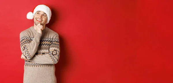 Bild eines glücklichen jungen Mannes mit Weihnachtsmütze und Weihnachtspullover, der etwas abbildet, über Neujahrsgeschenke nachdenkt und lächelt, in die linke obere Ecke blickt, vor rotem Hintergrund steht — Stockfoto