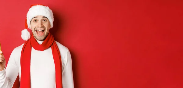 Noel, kış tatili ve kutlama kavramı. Yakışıklı adam yeni yılı kutluyor ve eğleniyor, maytap tutuyor ve gülümsüyor, Noel Baba şapkası takıyor, kırmızı arka planda duruyor. — Stok fotoğraf