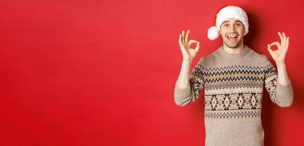 Портрет щасливого і задоволеного красивого чоловіка, одягненого в зимовий светр і капелюх Санти, демонструє гарні знаки, хвалить хорошу різдвяну вечірку, стоїть на червоному тлі — стокове фото