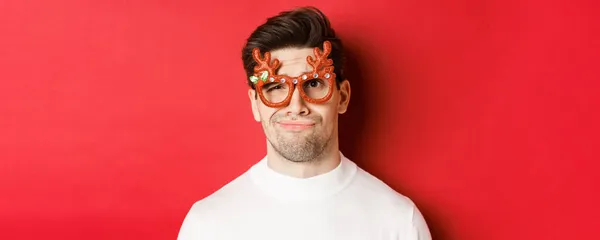 Käsite talvilomat, joulu ja juhla. Lähikuva skeptinen komea mies puolue lasit, etsivät epäilyttävä ja huvittunut, seisoo punaista taustaa vasten — kuvapankkivalokuva