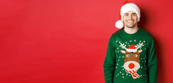 Concepto de vacaciones de invierno, Navidad y estilo de vida. Chico guapo con cerdas, con sombrero de santa y suéter verde, sonriente alegre, celebrando año nuevo, de pie sobre fondo rojo —  Fotos de Stock
