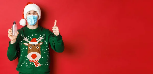 Концепція нового року, коронавірус та соціальне дистанціювання. Портрет веселого чоловіка в капелюсі Санта, різдвяний светр і медична маска, показує великий палець і рекомендує дезінфікуючий засіб для рук — стокове фото