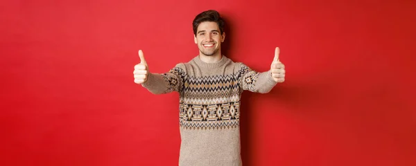 Imagem de belo homem caucasiano em camisola de Natal, mostrando polegares para cima na aprovação e sorrindo, desejando férias felizes, de pé sobre fundo vermelho — Fotografia de Stock