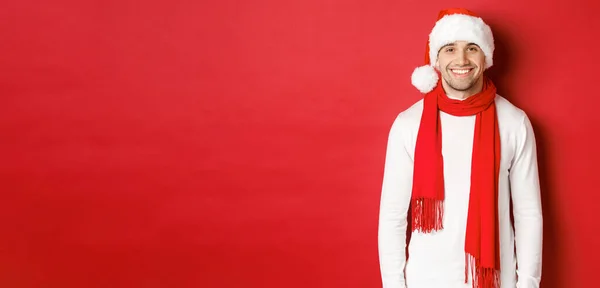 Mutlu beyaz adam Noel 'i kutluyor ve gülümsüyor, Noel Baba şapkası, atkı ve beyaz kazak giyiyor, yeni yılın tadını çıkarıyor, kırmızı arka planda duruyor. — Stok fotoğraf