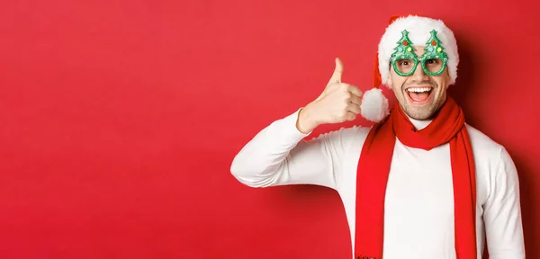 Noel, kış tatili ve kutlama kavramı. Noel Baba şapkası ve parti gözlüğü takmış neşeli bir adam. Başparmak kaldırıyor ve gülümsüyor. Kırmızı arka planda dikiliyor. — Stok fotoğraf