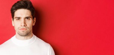 Kırmızı arka planda duran, beyaz kazaklı, düşünceli, ciddi görünümlü bir adamın yakın çekimi.