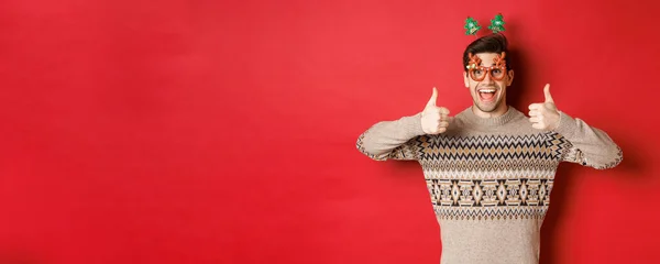 Retrato de homem satisfeito e feliz em camisola de Natal e óculos de festa, mostrando polegares para cima, desejando feliz ano novo, de pé sobre fundo vermelho — Fotografia de Stock