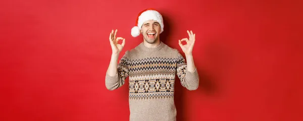 Bild eines fröhlichen attraktiven Kerls in Pullover und Weihnachtsmütze, der frohe Weihnachten wünscht, Zeichen zeigt und in die Kamera zwinkert, Neujahr feiert, vor rotem Hintergrund steht — Stockfoto