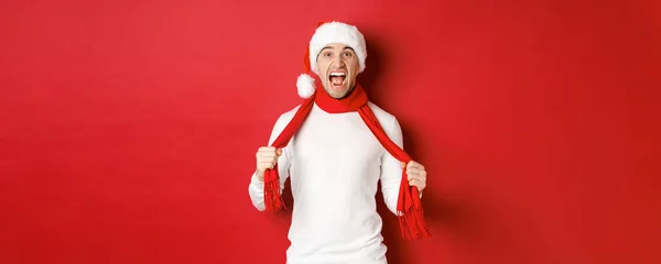 Зображення злий дорослий чоловік ненавидить Різдво, одягнений в шарф і Санта-капелюх, кричить засмучений, стоїть божевільним на червоному тлі — стокове фото