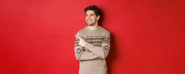 Retrato de hombre caucásico atractivo celebrando año nuevo, vacaciones de invierno, señalando el dedo y mirando a la izquierda con sonrisa complacida, mostrando el anuncio de Navidad, de pie sobre el fondo rojo —  Fotos de Stock