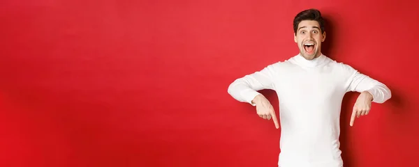Afbeelding van verbaasd knappe man in witte trui, reageren enthousiast op kerst advertentie, wijzend vingers naar beneden, tonen logo, staande over rode achtergrond — Stockfoto