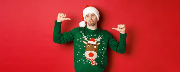 Imagen del joven guapo y confiado en suéter verde y sombrero de santa, apuntándose a sí mismo, celebrando la Navidad, de pie sobre el fondo rojo — Foto de Stock