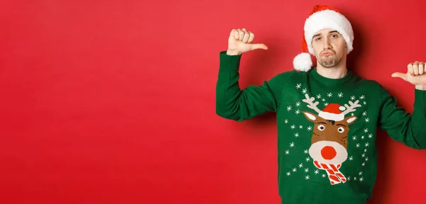 Beeld van knappe en zelfverzekerde jongeman in groene trui en kerstmuts, wijzend naar zichzelf, kerstviering, staande over rode achtergrond — Stockfoto