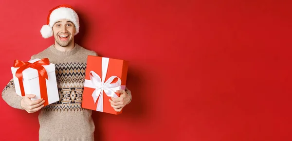 Bild eines fröhlichen attraktiven Kerls mit Weihnachtsgeschenken, der mit Weihnachtsmütze und Winterpullover vor rotem Hintergrund steht und glücklich lächelt — Stockfoto