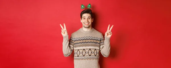 Concepto de vacaciones de invierno, Navidad y celebración. Imagen de chico guapo y tonto vestido para la fiesta de año nuevo, mostrando signos de paz y sonriendo, de pie sobre fondo rojo —  Fotos de Stock