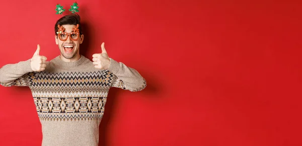 Retrato de homem satisfeito e feliz em camisola de Natal e óculos de festa, mostrando polegares para cima, desejando feliz ano novo, de pé sobre fundo vermelho — Fotografia de Stock