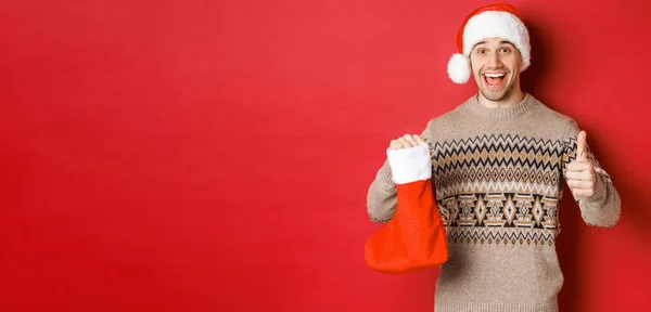 Conceito de férias de inverno, ano novo e celebração. Homem bonito alegre em chapéu de Papai Noel e suéter, mostrando Natal meia com doces e presentes, fazendo polegar-se — Fotografia de Stock