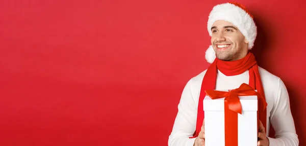 Conceito de férias de inverno, Natal e estilo de vida. Close-up de cara atraente em santa chapéu e cachecol, sorrindo e olhando para a esquerda enquanto segurando presente de ano novo, de pé sobre fundo vermelho — Fotografia de Stock