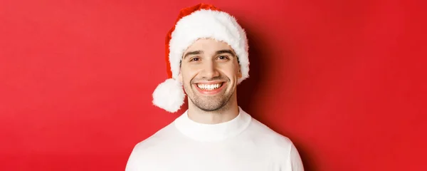 Primer plano del atractivo hombre sonriente en jersey blanco y sombrero de santa, luciendo feliz, disfrutando de las vacaciones de invierno, de pie sobre fondo rojo —  Fotos de Stock