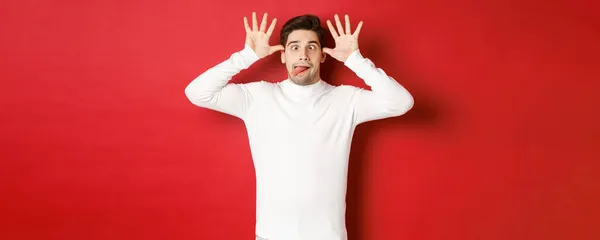 Porträt eines lustigen kaukasischen Typen, der Zunge zeigt und Gesichter macht, weißen Pullover trägt und vor rotem Hintergrund steht — Stockfoto