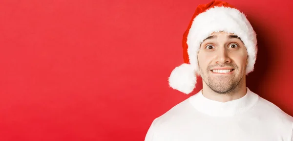 Primer plano de chico guapo en sombrero de santa, mordiendo el labio y mirando con tentación a algo que quiere, de pie sobre el fondo rojo —  Fotos de Stock