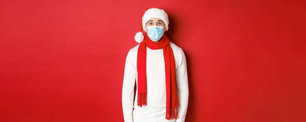 Neujahrskonzept, Coronavirus und Feiertage. Fröhlicher Mann, der Weihnachten und soziale Distanzierung feiert, mit medizinischer Maske, Weihnachtsmütze und Schal vor rotem Hintergrund — Stockfoto