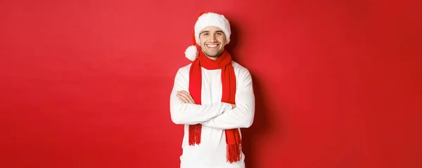 毛のあるハンサムな白人男性,サンタの帽子を身に着けています,スカーフと白いセーター,胸の上に腕を横断し、幸せな笑顔,赤の背景に立って — ストック写真