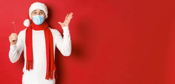 Concept de covid-19, Noël et vacances pendant la pandémie. Joyeux bel homme en masque médical et chapeau de Père Noël, célébrant le nouvel an avec scintillant, regardant excité, fond rouge — Photo
