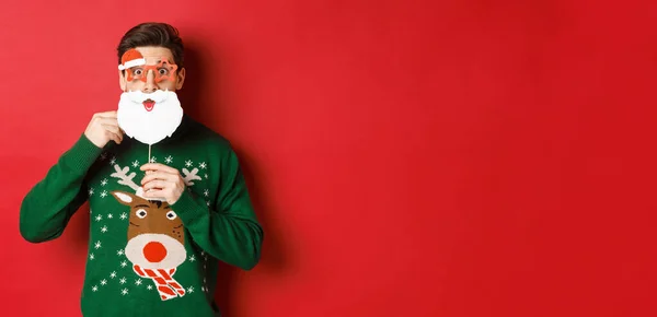 Retrato de homem surpreso em camisola de Natal verde, segurando engraçado máscara de Papai Noel, celebrando o ano novo, de pé sobre fundo vermelho — Fotografia de Stock