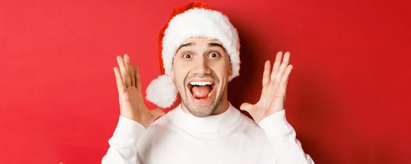 Zbliżenie szczęśliwego młodzieńca w kapeluszu Mikołaja, wielkie ogłoszenie świąteczne, uśmiechnięty zdumiony, stojący na czerwonym tle — Zdjęcie stockowe