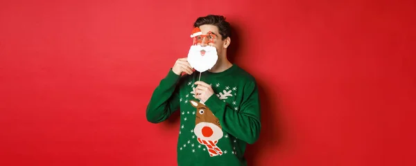 Porträtt av överraskad man i grön jul tröja, håller rolig Santa claus mask, firar nytt år, tittar vänster förvånad, slipning över röd bakgrund — Stockfoto