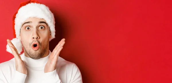 Großaufnahme eines erstaunten gutaussehenden Mannes mit Weihnachtsmannmütze und weißem Pullover, der die Hände hebt und beeindruckt wirkt, vor rotem Hintergrund stehend — Stockfoto
